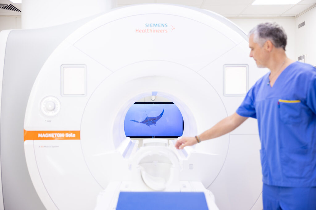 Ein Mitarbeiter der Univ.-Klinik für Radiologie zeigt ein MRT-Gerät mit Entertainmentsystem. Auf einem Monitor ist ein Video mit einem Rochen zu sehen.
