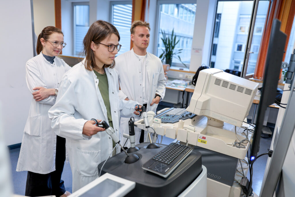 Die Studierende Julia von Fugler übt eine Laparoskopie, indem sie zwei Steuerungselemente vor einem Monitor bedient, beobachtet von ihrem Studienkollegen Felix Hirtenlehner und der Ärztin Irina Tsibulak