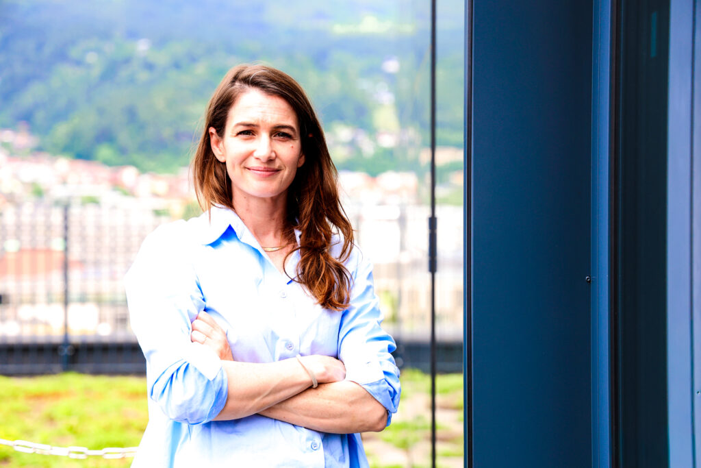 Die Alzheimer-Forscherin Lindsay Lohan steht vor einem Fenster an der Medizinischen Universität Innsbruck. Im Hintergrund sieht man die Berge bei sommerlichem Wetter.